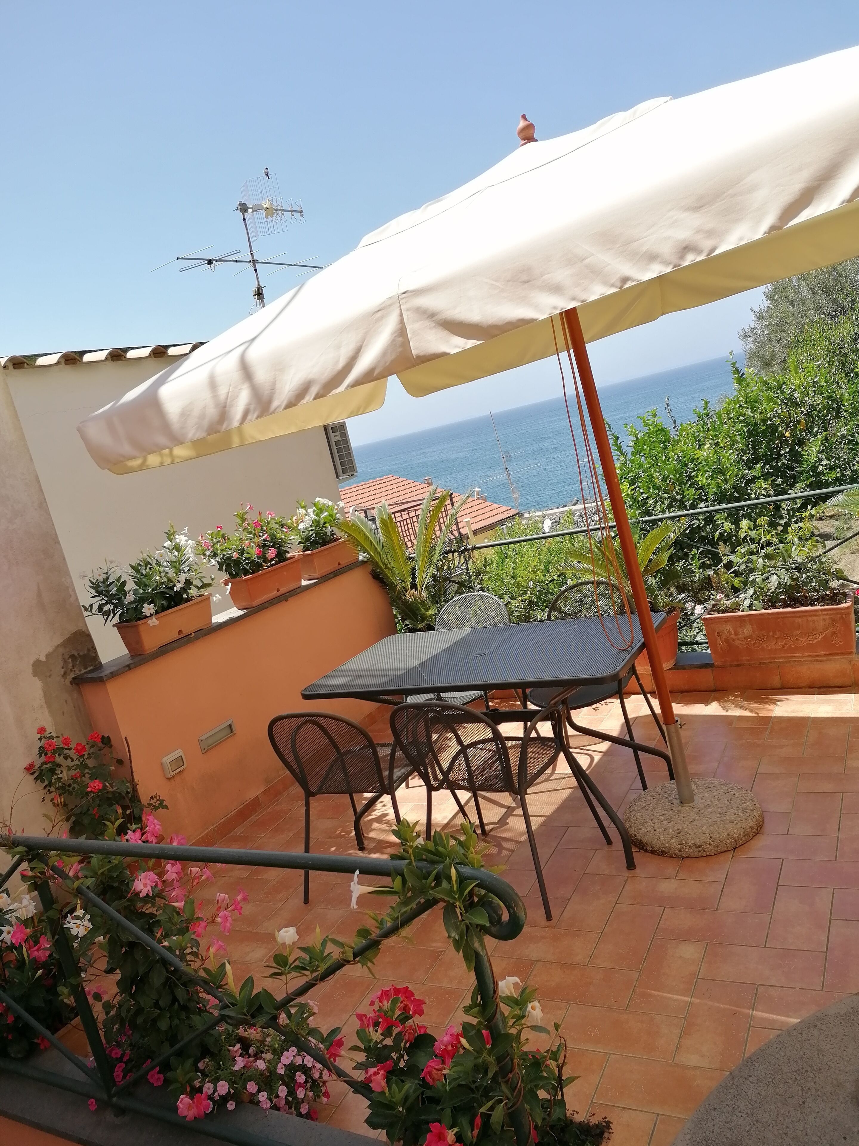 A patio with a table and chairs, a large umbrella, potted plants, and a view of the sea.