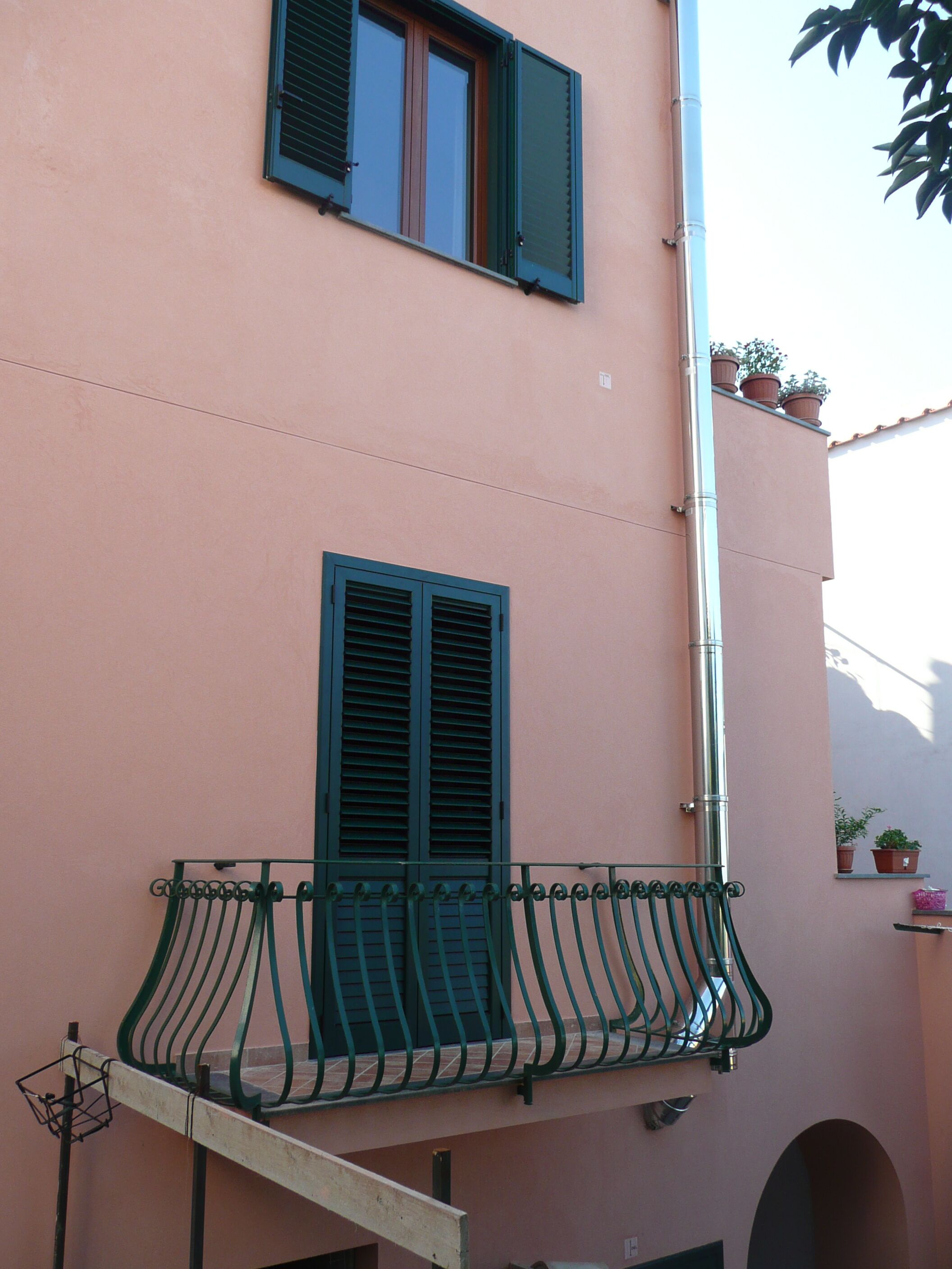 Un edificio con fachada rosa, un balcón con barandillas de metal verde y dos ventanas con persianas verdes.