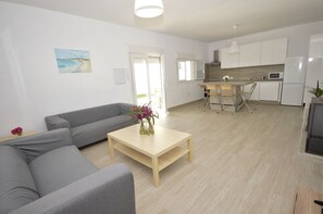 Ein modernes Wohnzimmer mit einem grauen Sofa, einem Holztisch mit Blumenvase und einer Küchenzeile mit weißen Schränken und Geräten.