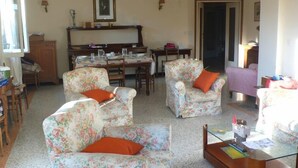 Uma sala de estar com poltronas estampadas, uma mesa de centro, uma mesa de jantar posta para quatro pessoas e um piano ao fundo.