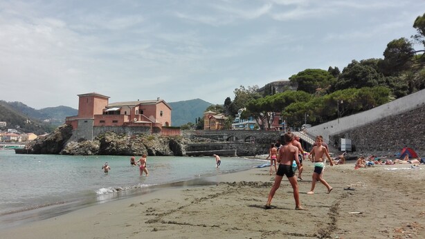Uma praia com pessoas nadando e caminhando, um muro de pedra e edifícios ao fundo.