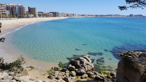 A coastal beach with clear water, a rocky shoreline, and a cityscape in the background.