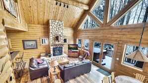 Ein Blockhaus-Interieur mit Kamin aus Naturstein, Holzmöbeln und großen Fenstern mit Blick auf eine verschneite Landschaft.