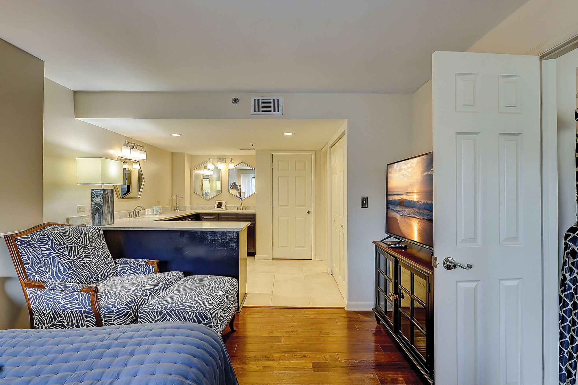 A hotel room with a bed, a seating area with patterned chairs, a kitchenette, a bathroom with mirrors, and a TV displaying a beach scene.