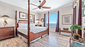 Une chambre spacieuse dotée d’un lit à baldaquin, de parquet, d’un ventilateur de plafond et d’une grande fenêtre offrant une vue sur l’océan.