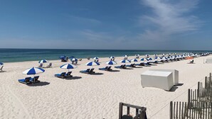 Une plage bordée de rangées de parasols bleus et blancs et de chaises longues, un bâtiment blanc et une clôture en bois.