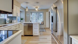 Une cuisine moderne dotée d’un îlot central, d’appareils électroménagers en acier inoxydable, de luminaires suspendus et offrant une vue sur l’extérieur grâce à une fenêtre.
