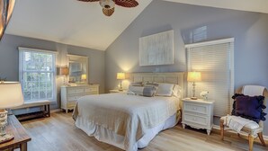 Une chambre à coucher avec un grand lit, deux tables de chevet avec des lampes, un ventilateur de plafond et une fenêtre avec des stores.
