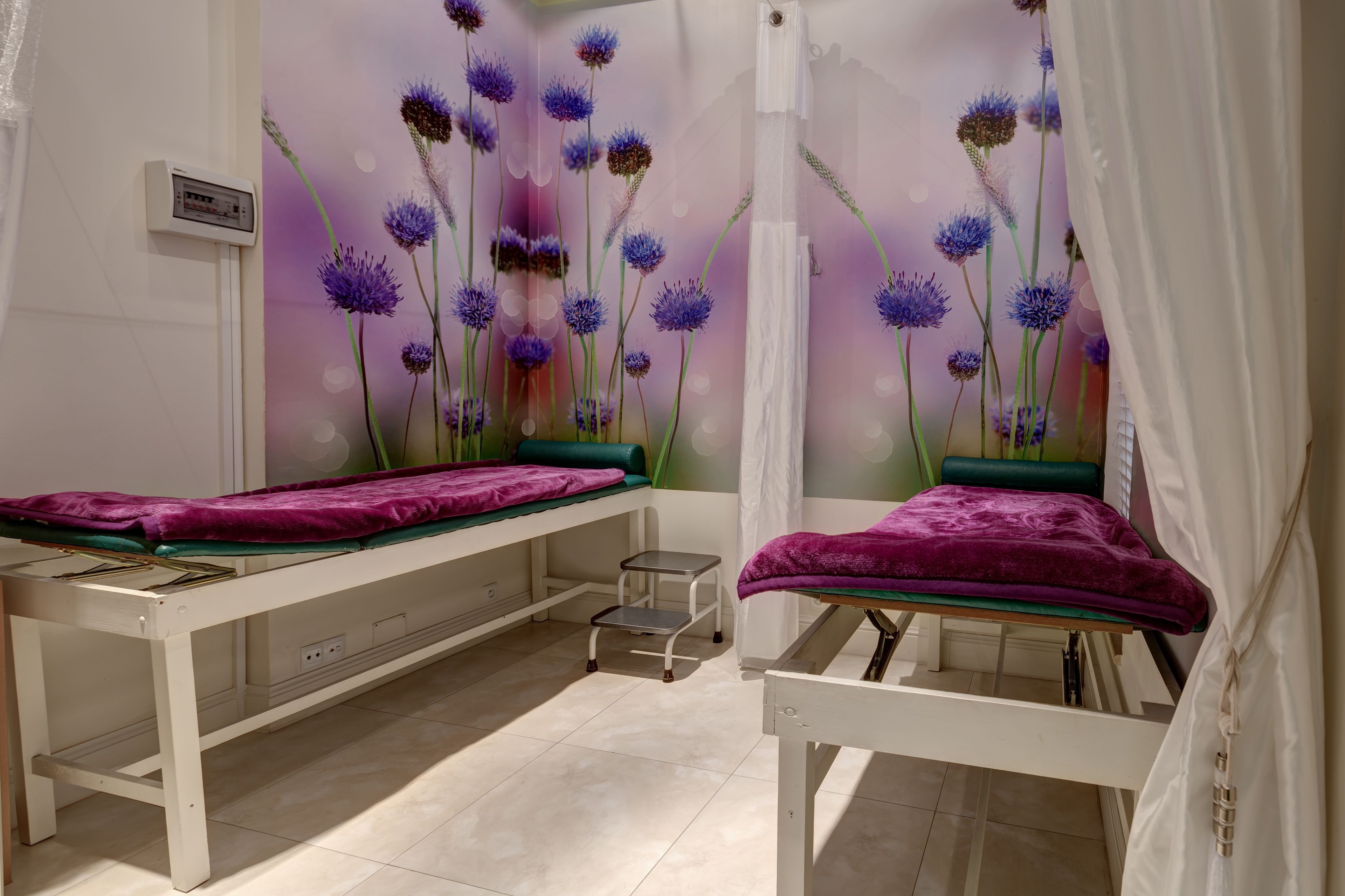 A medical examination room with two examination tables, each with a purple cover, and a wall mural of purple flowers.