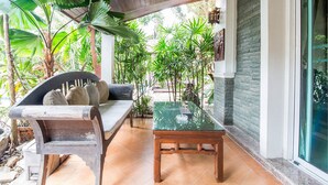 Un espacio de estar al aire libre techado con un banco de madera y una mesa de vidrio, rodeado de exuberante follaje tropical.