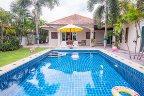 A swimming pool with a tiled deck, a covered seating area with chairs and an umbrella, and a house with a tiled roof in the background. - Baan Sod Sai HuaHin Pool Villa (Hua Hin)