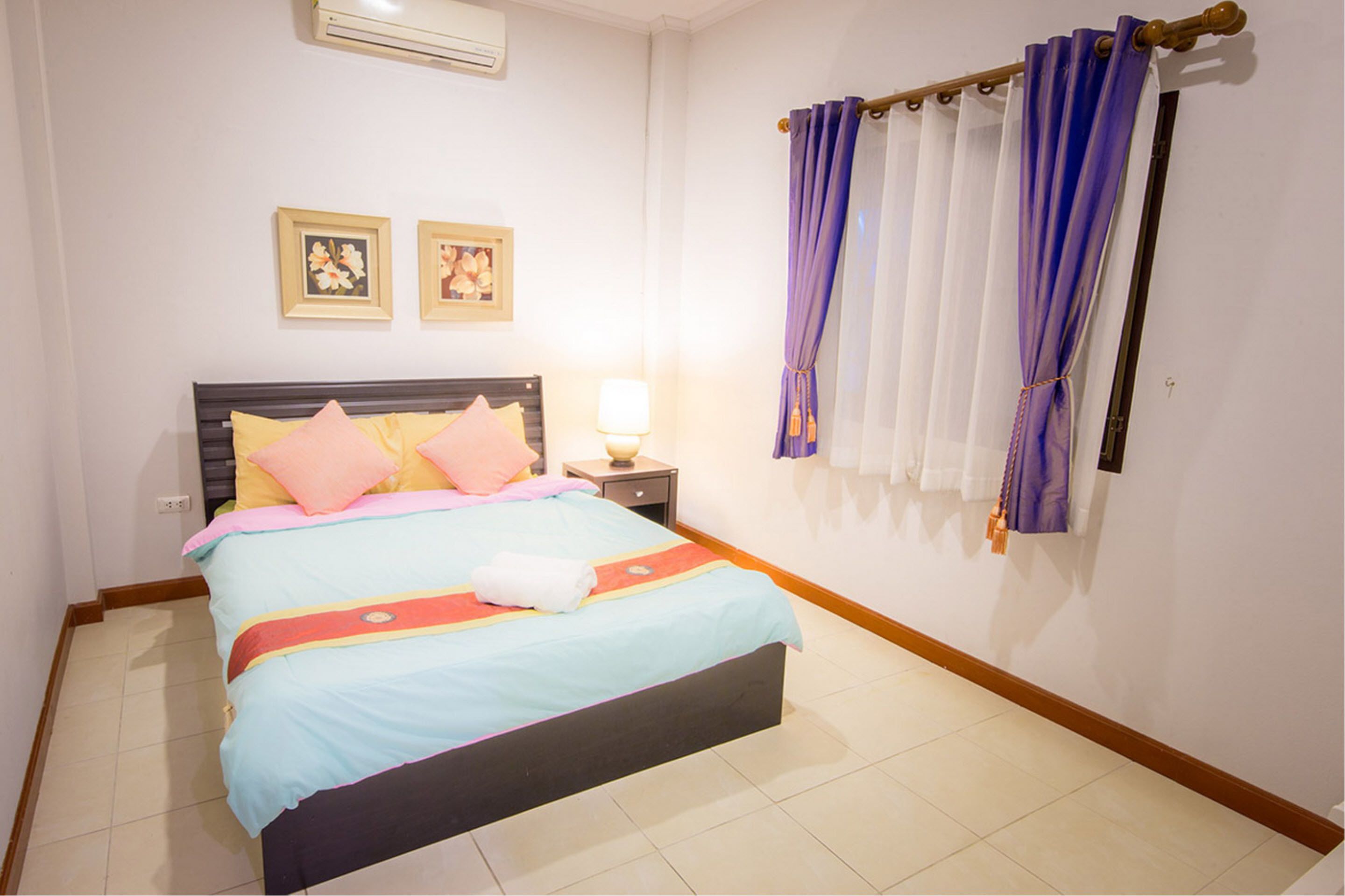 A neatly made bed with a blue and pink bedspread, a bedside table with a lamp, and two framed pictures on the wall.