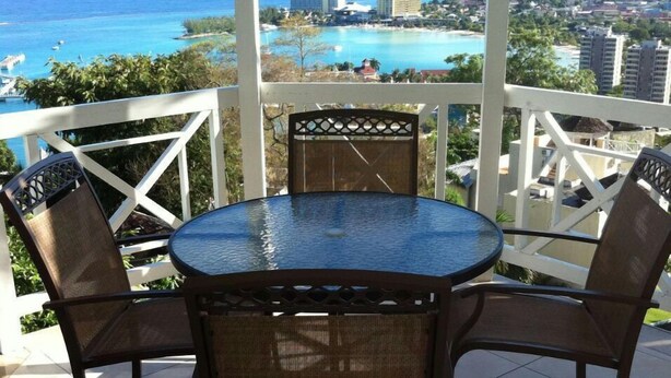 A round glass table with four chairs, a white railing, and a view of the ocean and buildings. - Columbus Heights Baywatch (Ocho Rios)