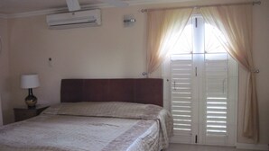 A bedroom with a bed, an air conditioner, a nightstand with a lamp, and a window with curtains. - Columbus Heights Baywatch (Ocho Rios)