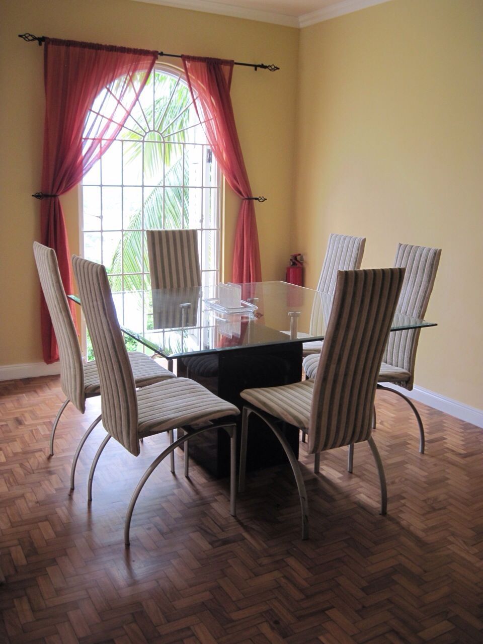 A dining area with a glass table surrounded by four striped chairs, a window with red curtains, and a herringbone wooden floor.