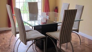 A dining area with a glass table surrounded by six striped chairs, a window with red curtains, and a wooden herringbone floor. - Columbus Heights Baywatch (Ocho Rios)