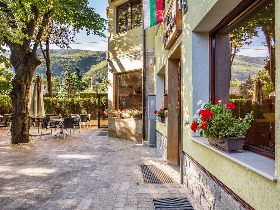 Un espace café en plein air avec des tables et des chaises, un arbre et un bâtiment dont la fenêtre est ornée de fleurs rouges.
