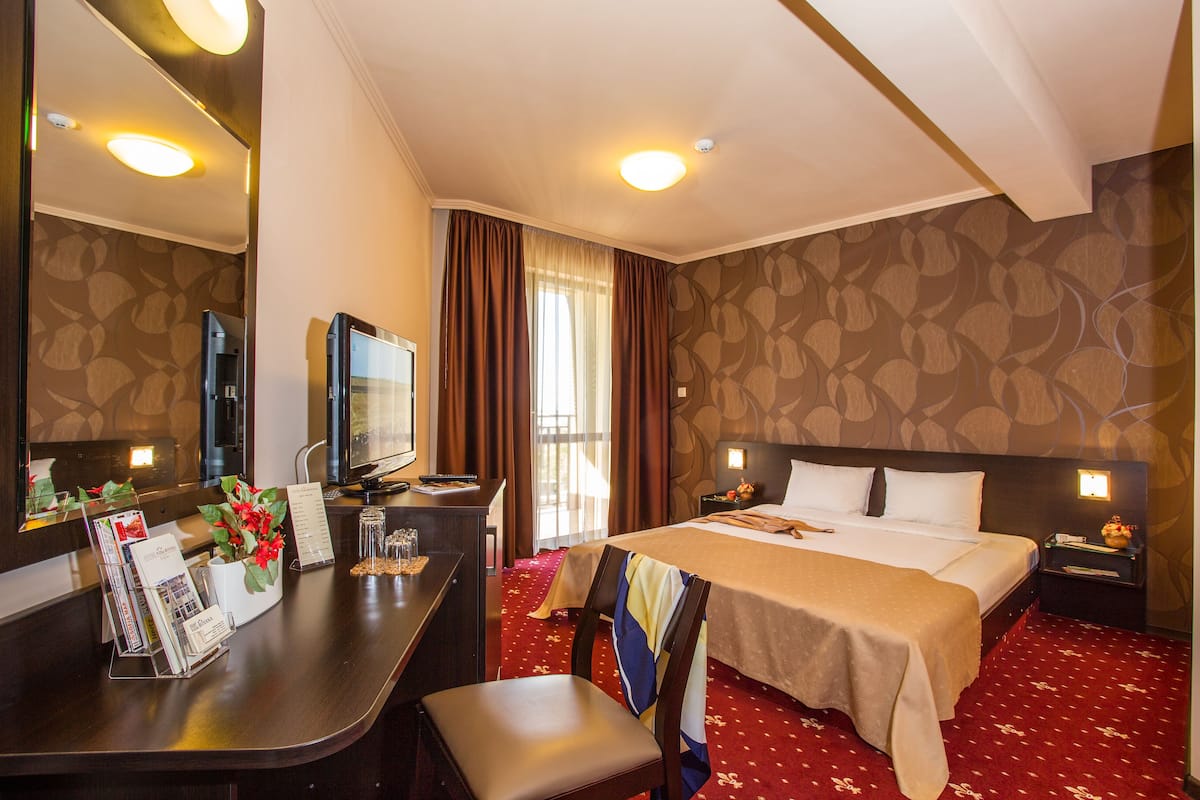 A hotel room with a bed, a desk with a computer, a chair, a TV, and a large mirror.