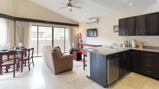 A modern kitchen with dark cabinets, a built-in dishwasher, and a large island. There is a dining area with a table and chairs, and a sofa. - The Cove 4 at Playa Ocotal (Sardinal)