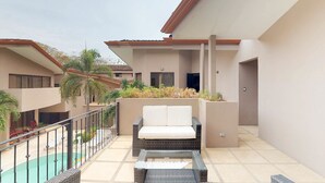 Un balcón con un sofá de mimbre y una mesa de centro de vidrio, una piscina y un edificio con techo de tejas y paredes color beige.