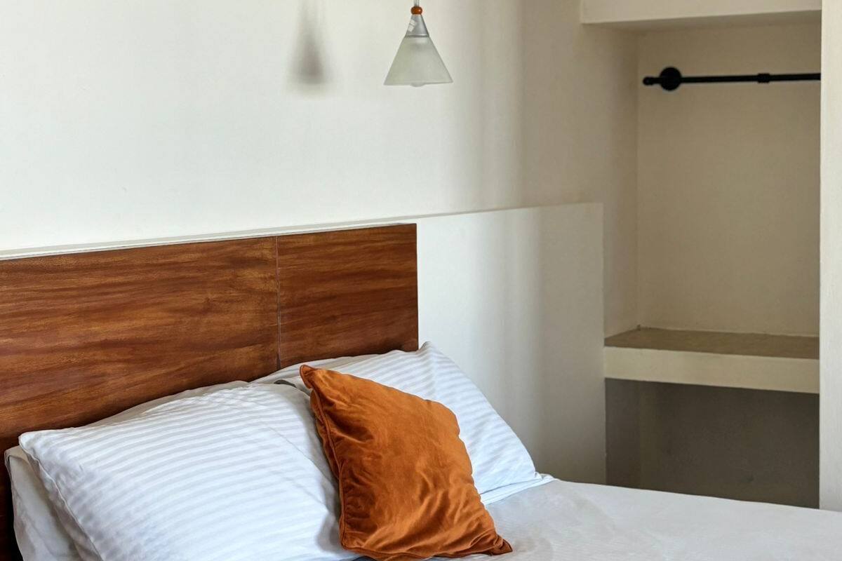 A bedroom with a wooden headboard, a bed with white bedding and a brown pillow, a hanging light fixture, and a built-in air conditioning unit.