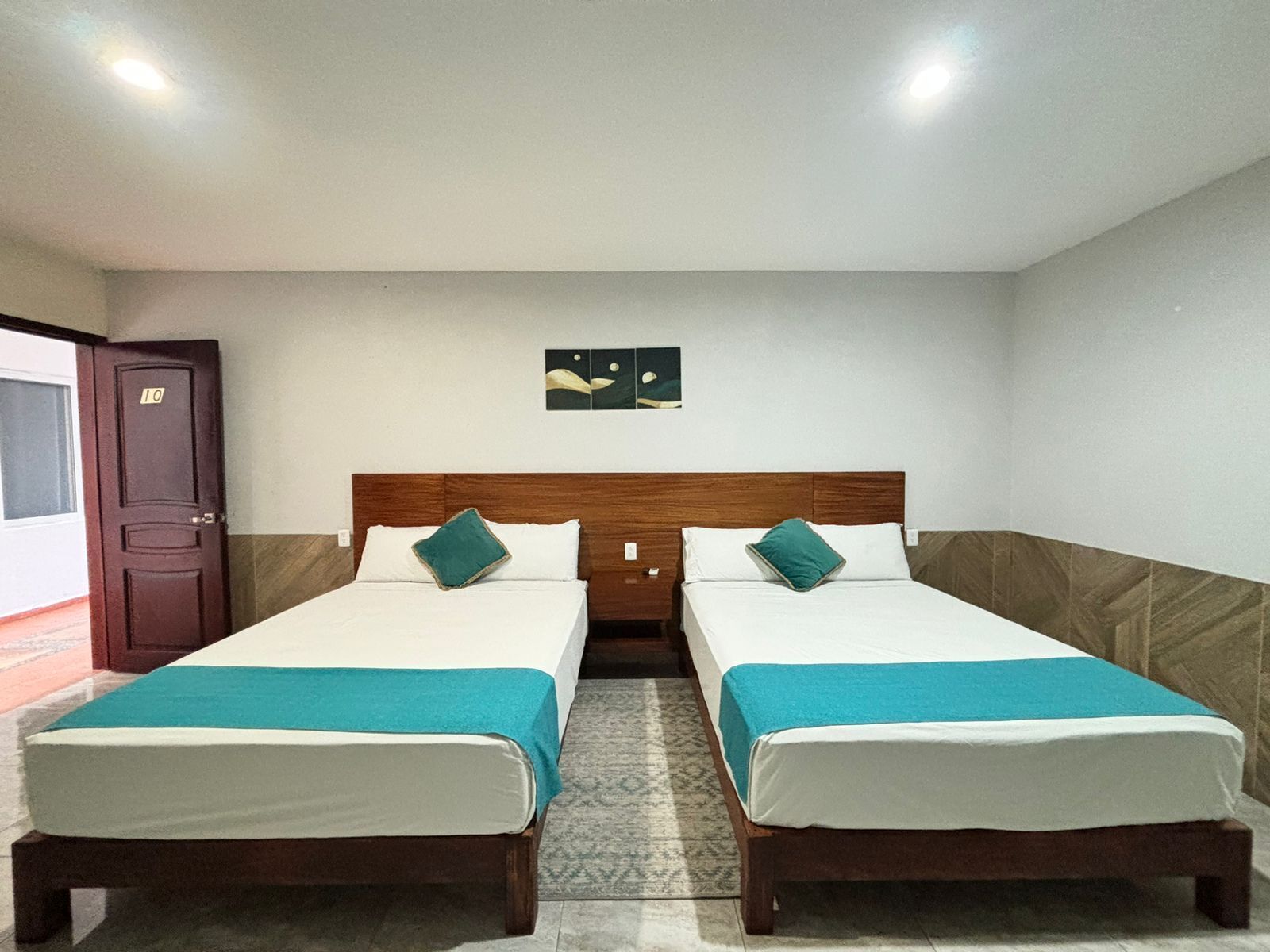 A hotel room with two single beds, a wooden headboard, a small wall art piece, and a door with a number.
