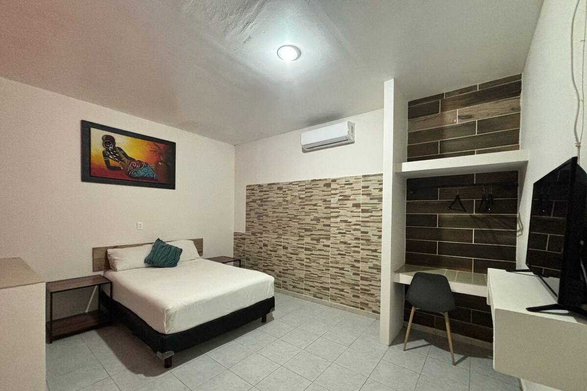 A bedroom with a bed, a desk, a chair, a painting, an air conditioner, and a brick-patterned wall.