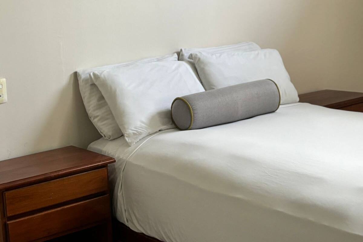 A neatly made bed with white linens and two pillows, a wooden nightstand with a drawer, and a wall with a grey and white patterned design.