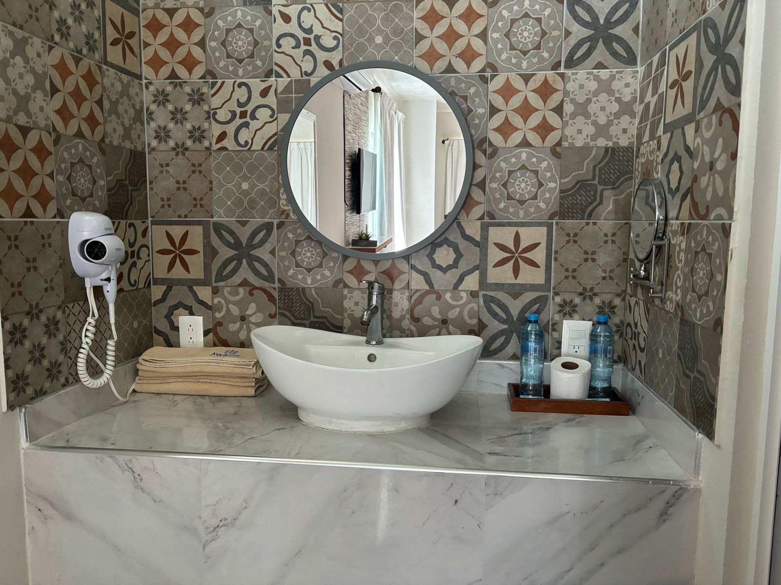 Une salle de bain avec un lavabo blanc, un miroir rond, un sèche-cheveux et des carreaux à motifs.