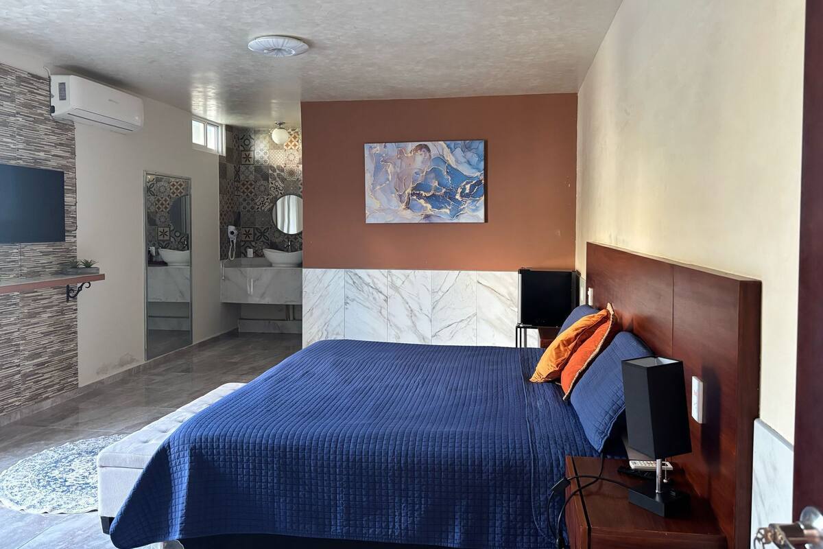 A hotel room with a bed, a desk with a computer, a TV, and a bathroom with a mirror and sink.