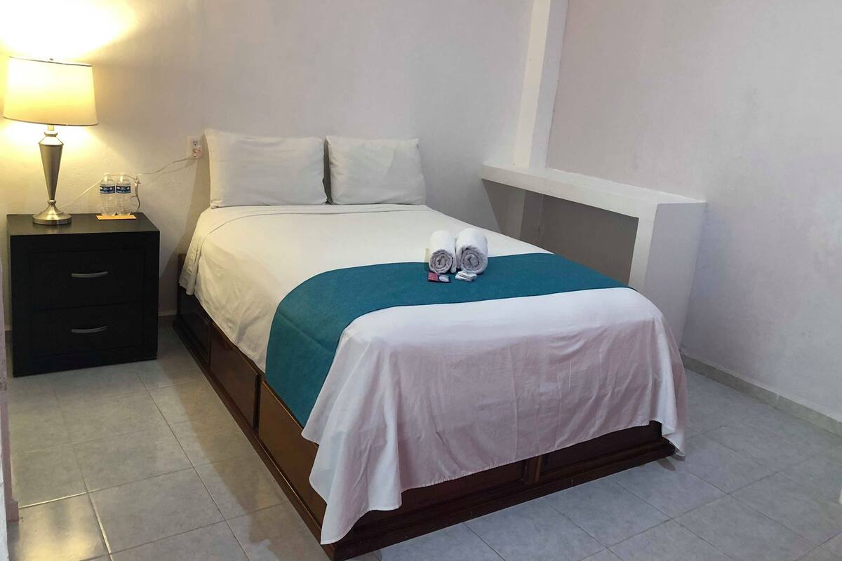 A hotel room with a bed, a nightstand with a lamp, and a water bottle.
