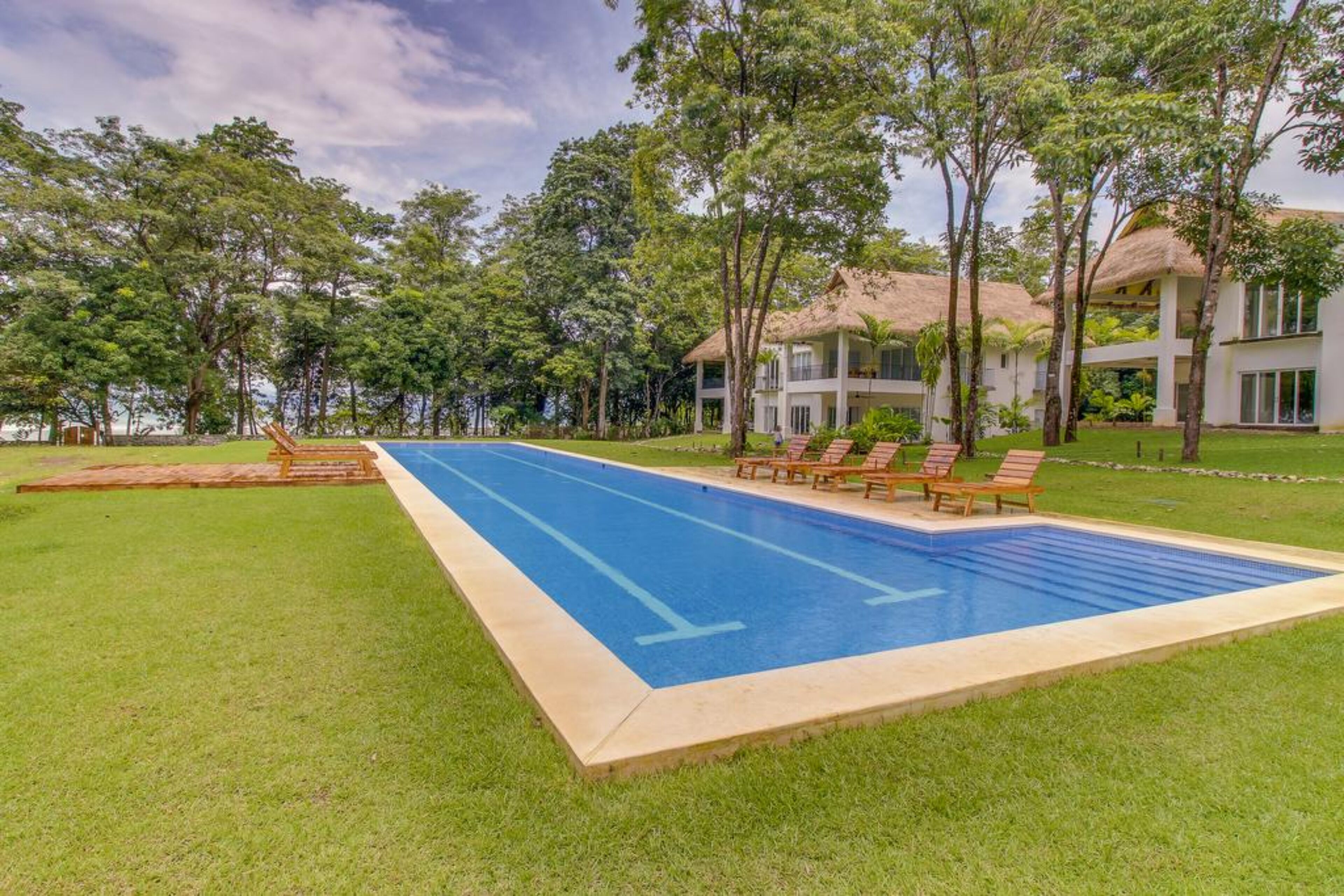 Una piscina con sillas de descanso, rodeada de árboles y un edificio con techo de paja.