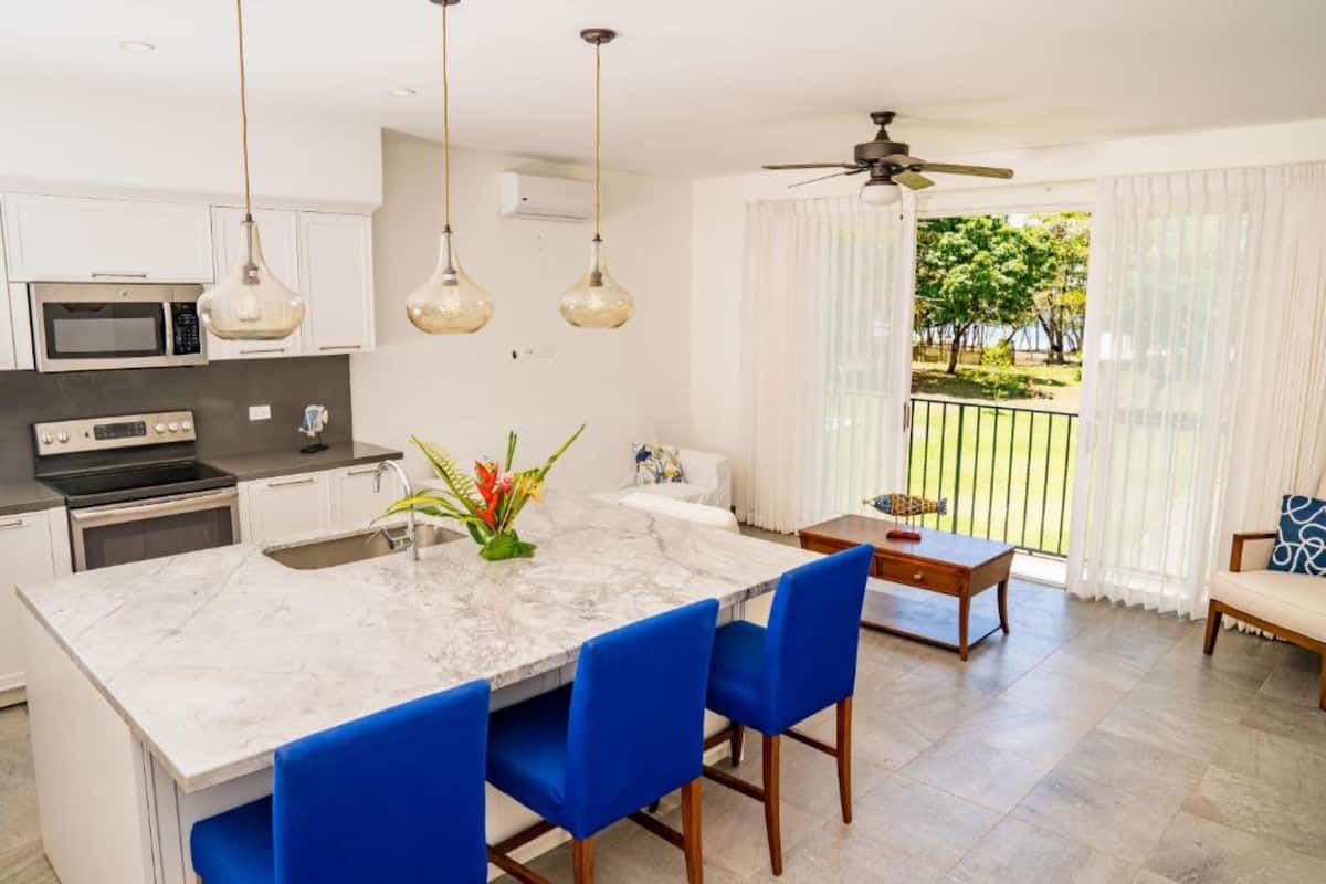 A modern kitchen with white cabinets, a marble island, and blue chairs. There is a dining area with a table and chairs, and a balcony with a view of greenery.
