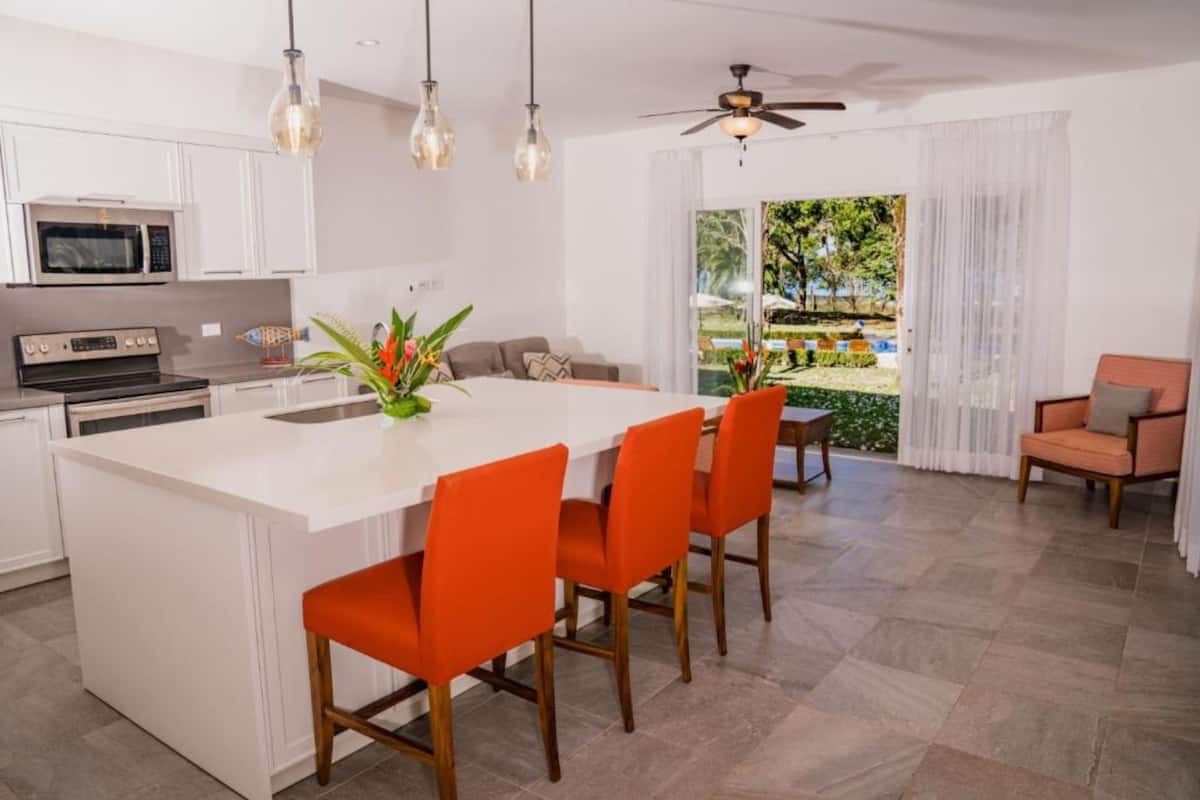 A modern kitchen with white cabinetry, a central island with orange chairs, a dining area with a table and chairs, and a view of a garden through a sliding door.