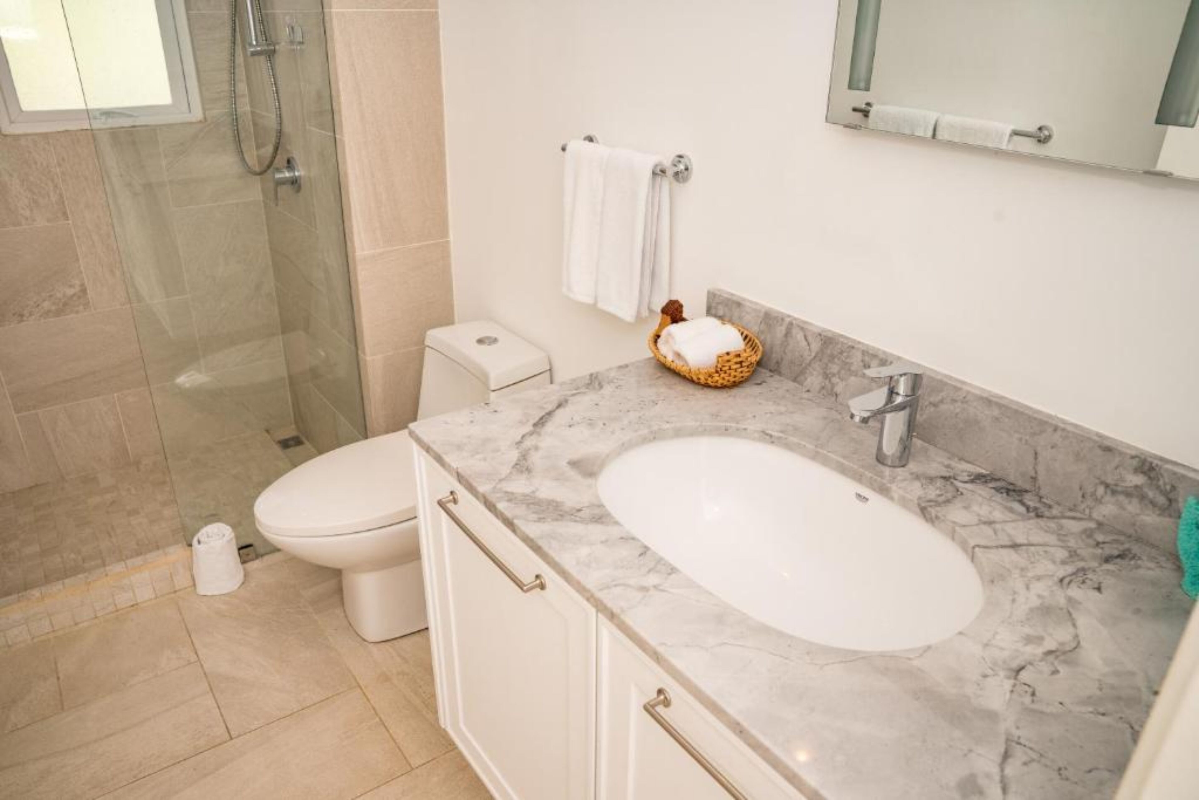 Un baño con cabina de ducha de vidrio, inodoro, lavabo con encimera de mármol y una cesta con toallas.