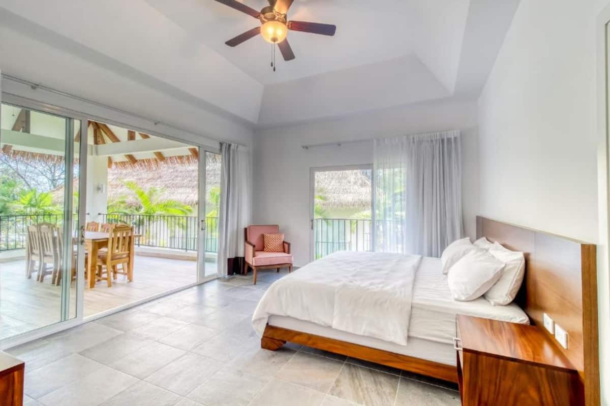 A bedroom with a bed, a ceiling fan, a window with curtains, and a balcony with a table and chairs.
