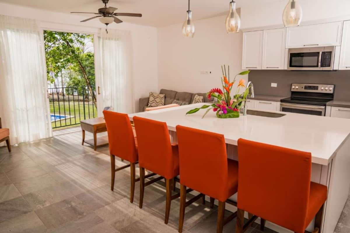 A modern kitchen with white cabinets, a central island with orange chairs, and a dining area with a view of a pool.