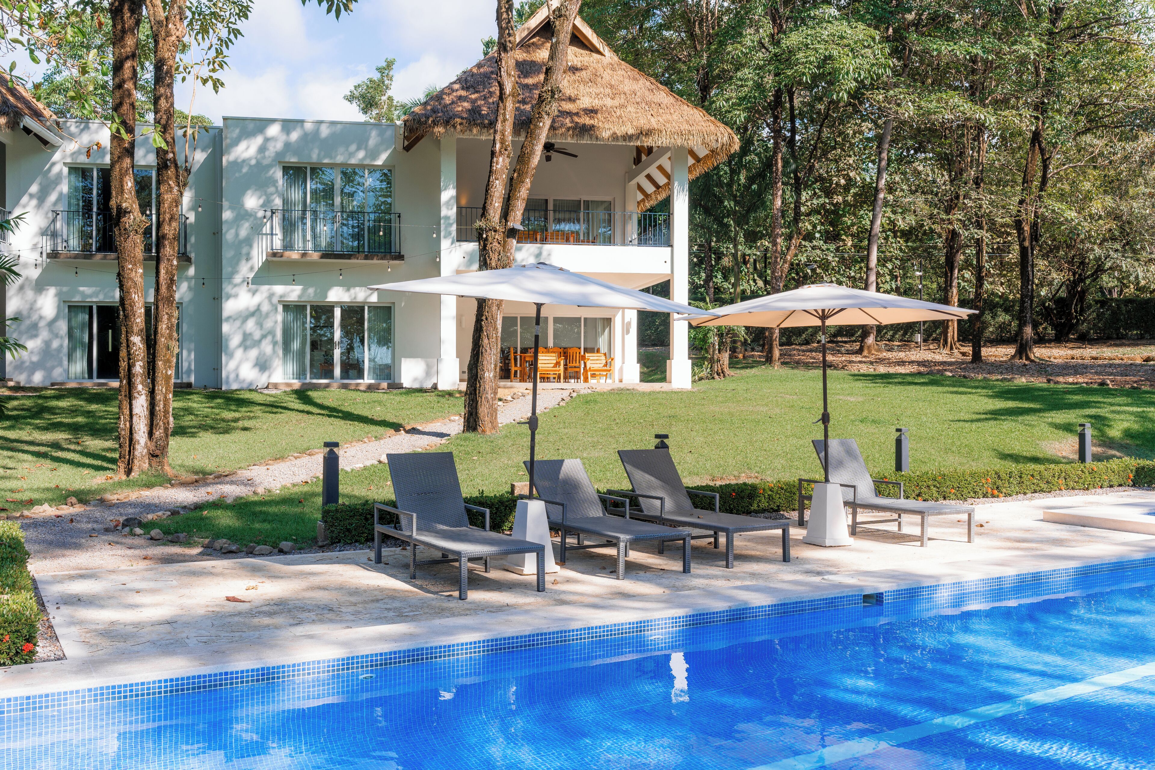A modern two-story house with a swimming pool, outdoor seating, and a thatched-roof structure.