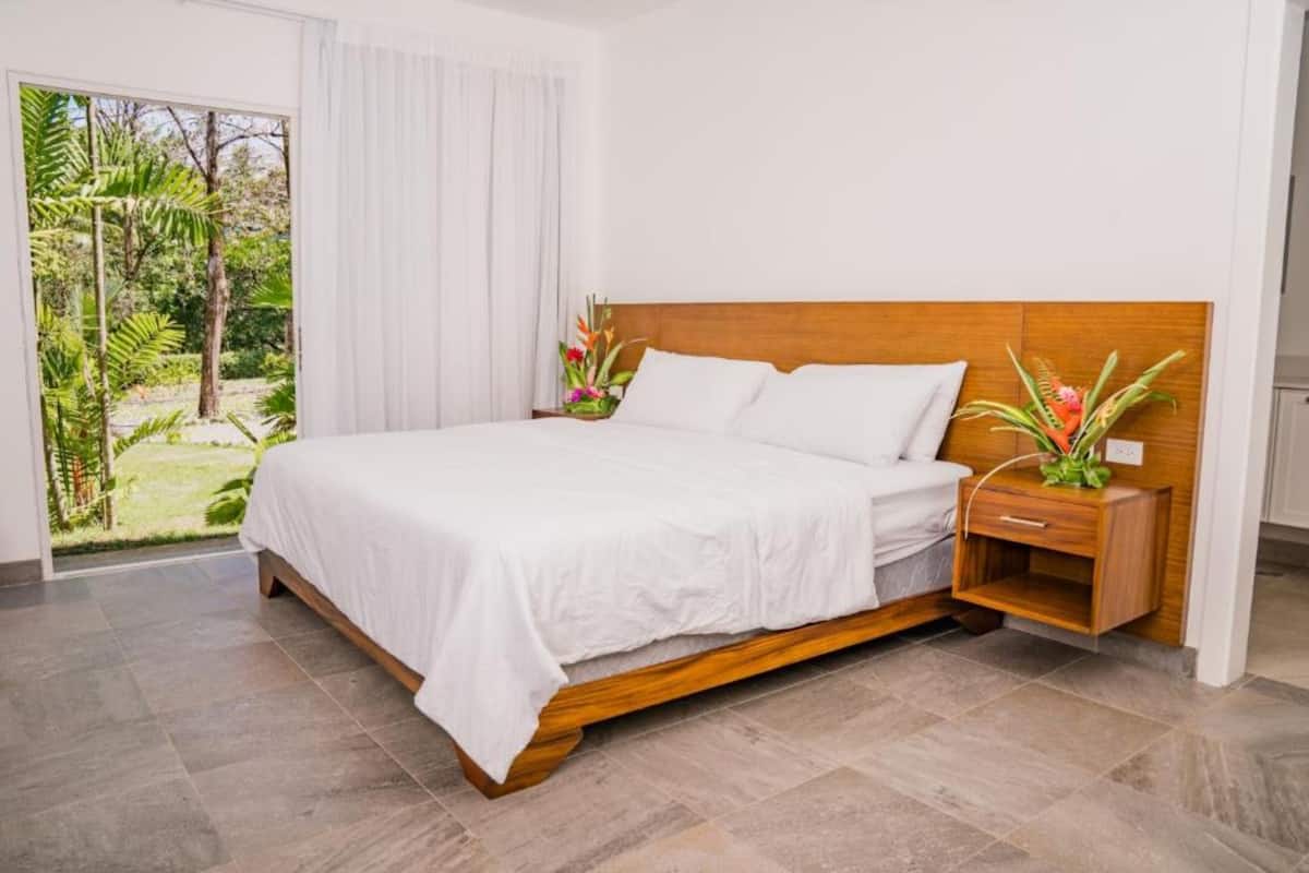A bedroom with a wooden bed frame, white bedding, a nightstand with a vase of flowers, and a view of greenery through a large window.