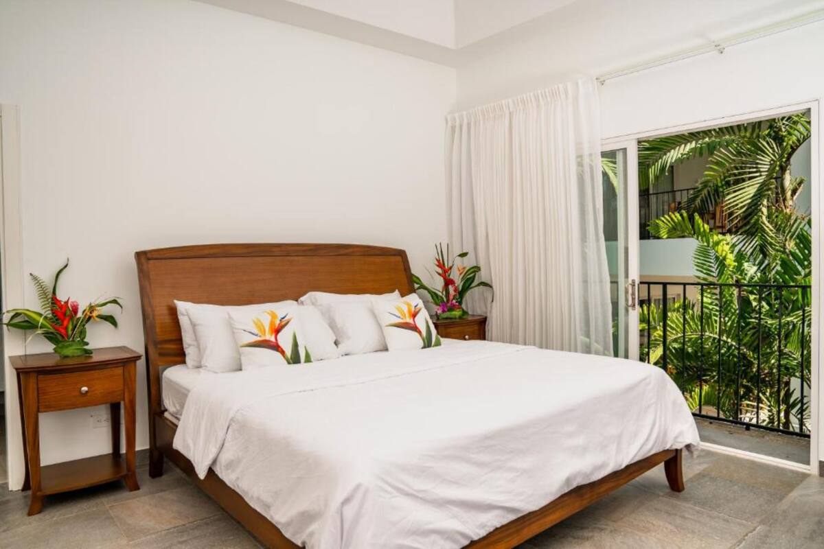 A bedroom with a wooden bed, white bedding, two nightstands with flower vases, and a balcony with plants.