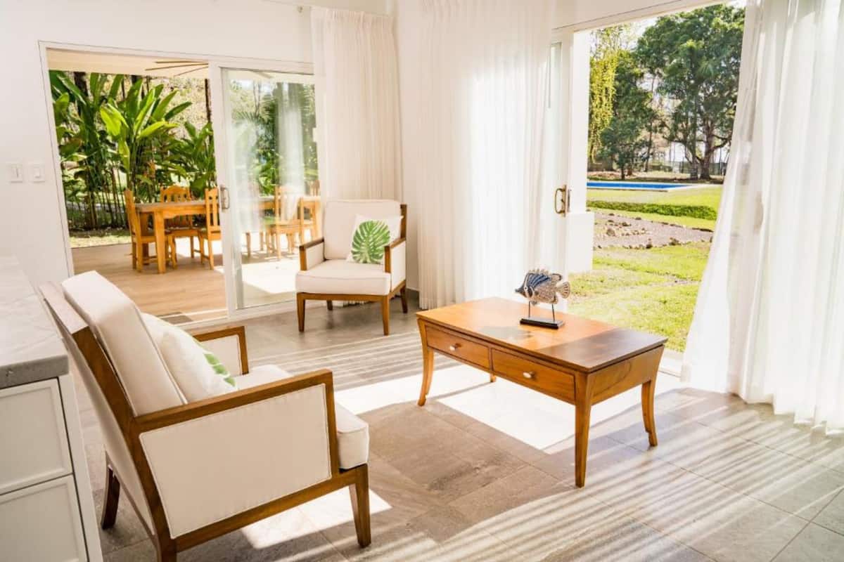 A spacious living area with a sofa, a wooden coffee table, and a chair with a green leaf pillow. There is a glass door leading to an outdoor garden area.