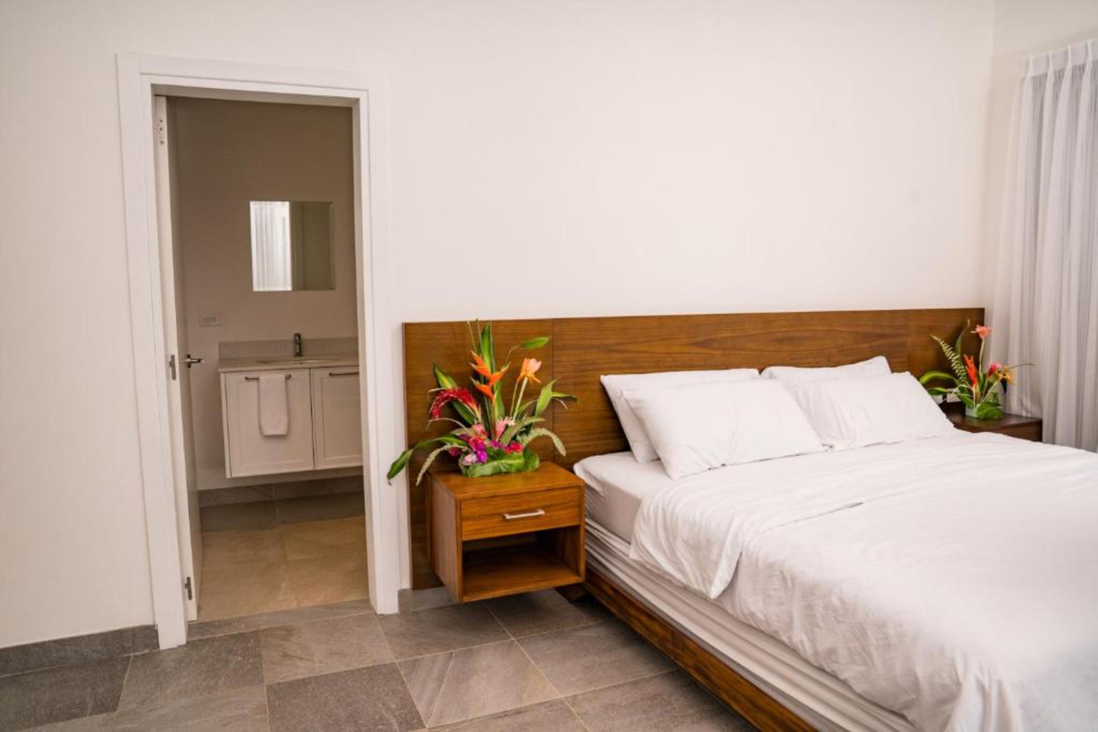 Ein Hotelzimmer mit hölzernem Kopfteil, weißer Bettwäsche, einem Nachttisch mit Blumenvase und Blick ins Badezimmer durch eine geöffnete Tür.