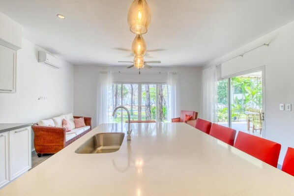 A modern kitchen with a large island, a dining area with red chairs, and a view of greenery outside.