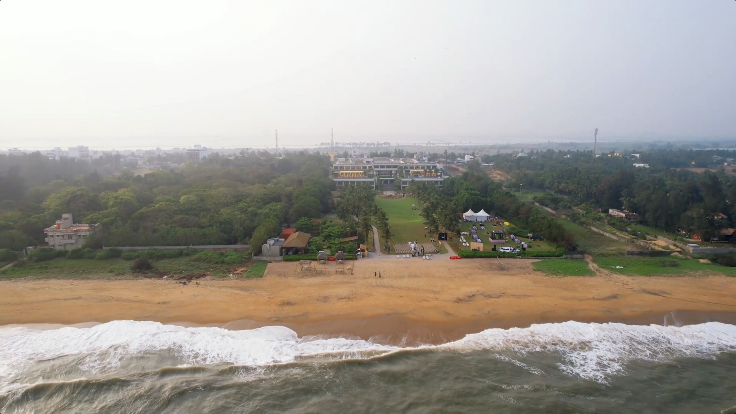 An aerial view of a coastal area with a beach, a large building complex, and surrounding greenery.