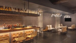 Zona de bar con mostrador de madera, taburetes y una variedad de botellas. Hay luces colgantes y un área de estar con sillas y un sofá.