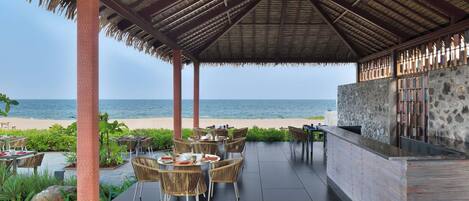 An outdoor seating area with tables and chairs, a stone wall, and a view of the beach.