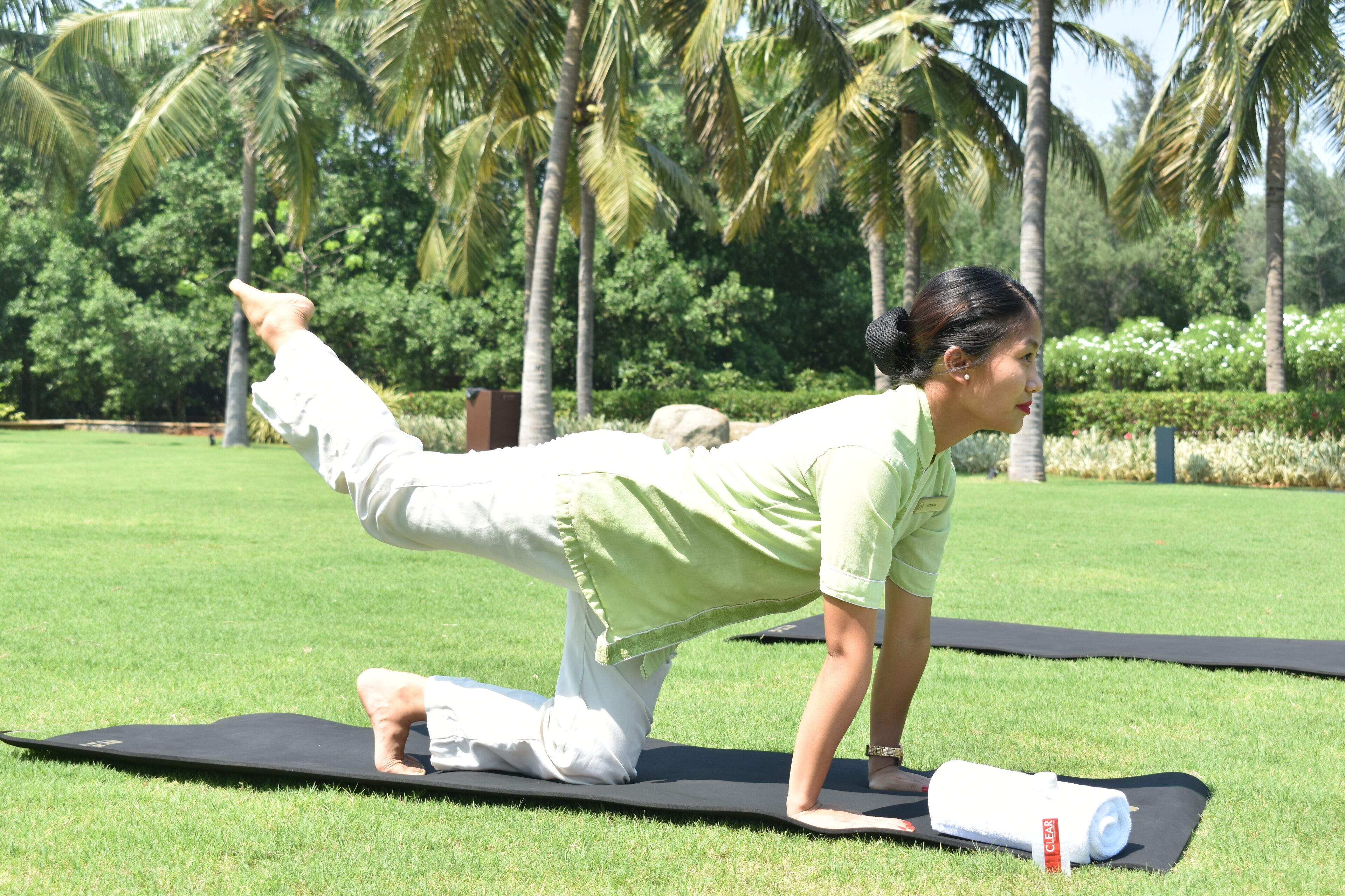 A person performing a yoga pose on a mat in a grassy area with palm trees in the background.