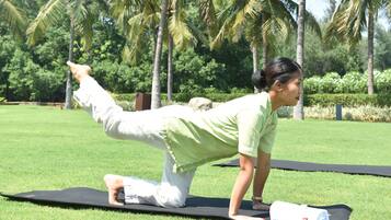 A person performing a yoga pose on a mat in a grassy area with palm trees in the background.