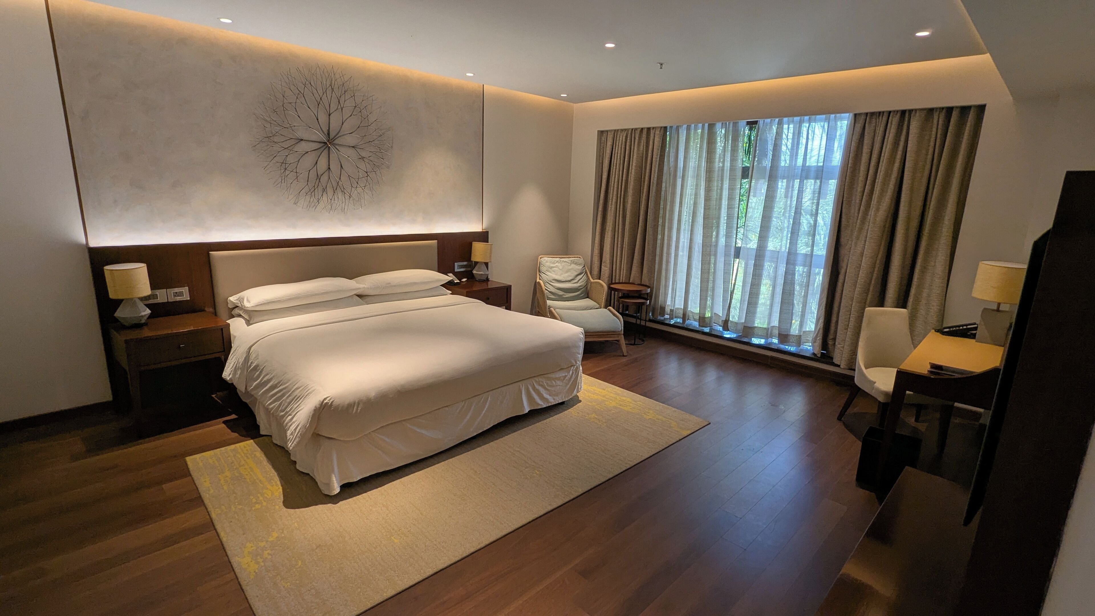 A modern hotel bedroom with a large bed, a wall-mounted clock, and a view of greenery through the window.