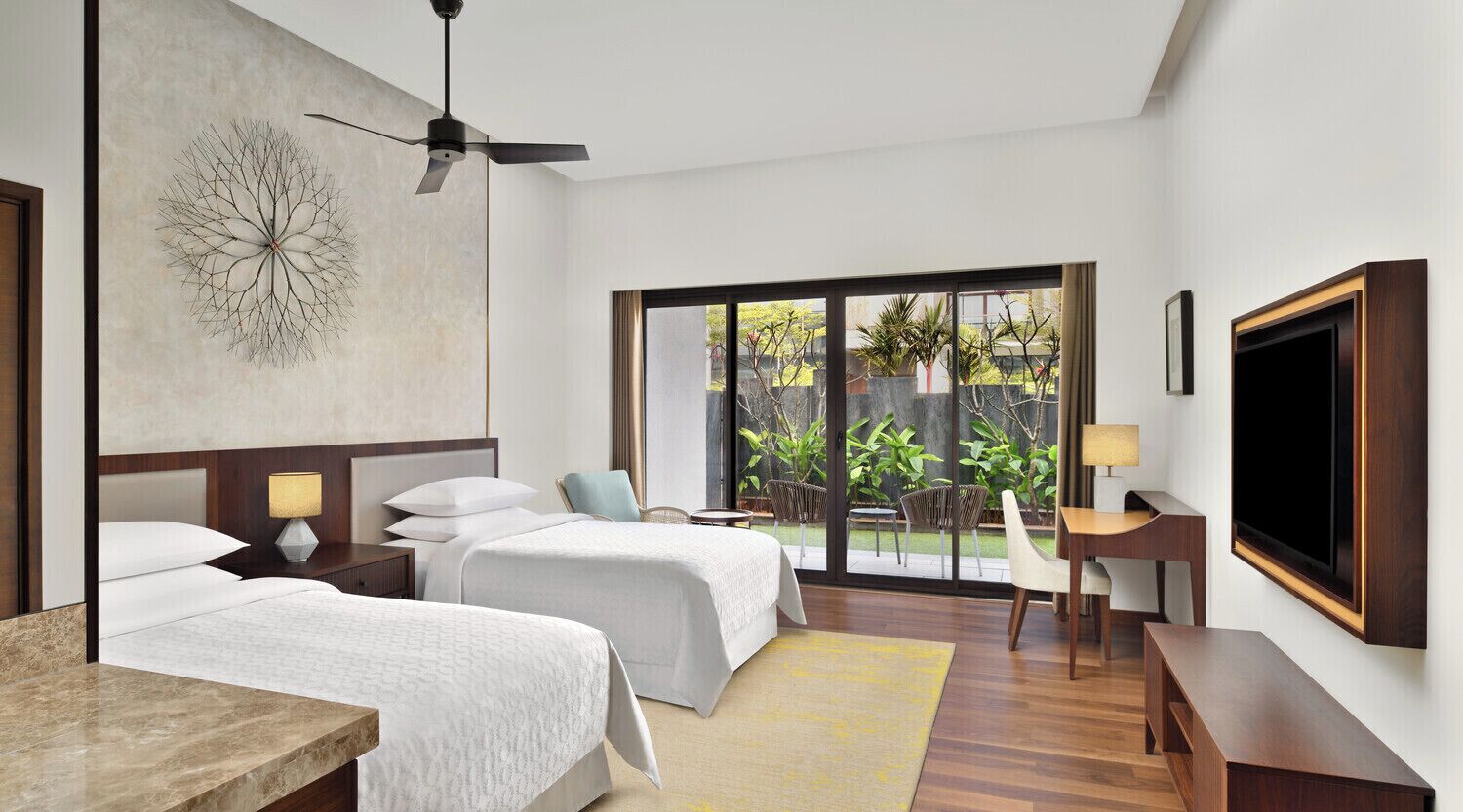 A hotel room with two beds, a desk, a TV, and a balcony with plants.